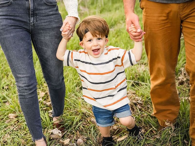 Young boy walking outside holding his parents hands.